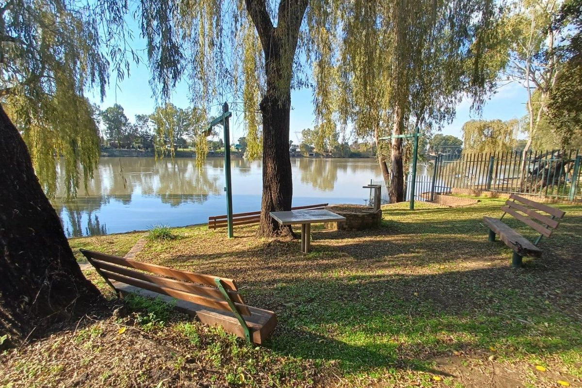 A scenic riverside garden area featuring mature willow trees, built-in benches, and a small table, offering direct access to the water.