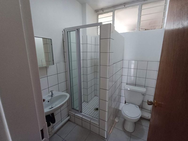 A functional bathroom featuring a shower enclosure, pedestal basin, and toilet with white tiled finishes and natural light from high-level louvre windows.