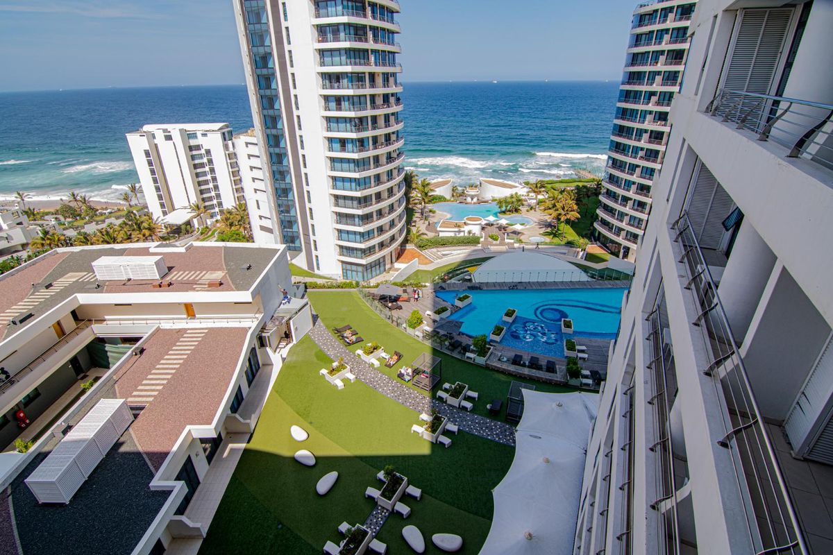 Elevated aerial view overlooking the apartment complex grounds, featuring a swimming pool, landscaped garden areas, and direct ocean views.