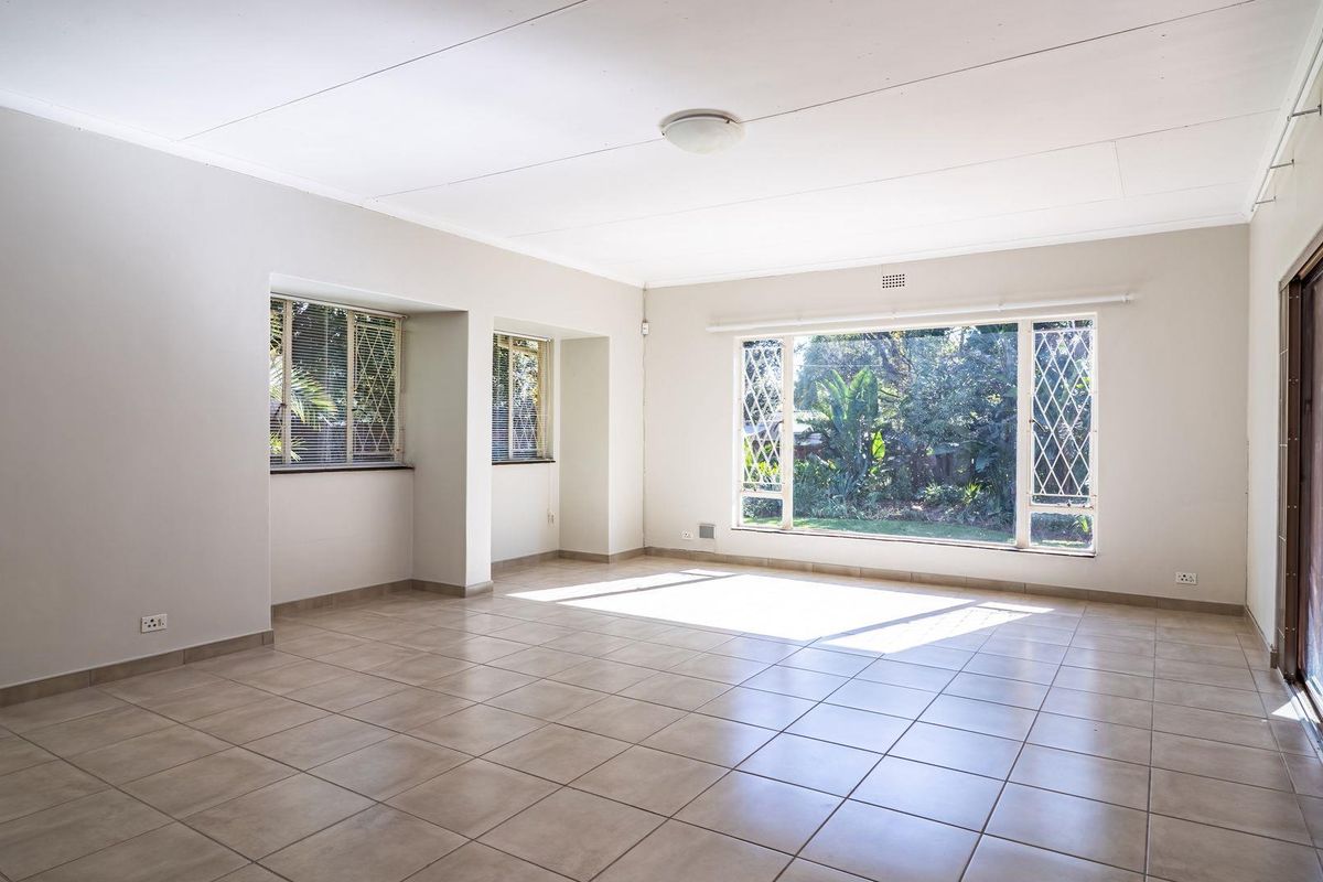 A spacious, sunlit living room featuring neutral ceramic floor tiles, large windows with garden views, and sliding glass doors leading to the outdoor area.