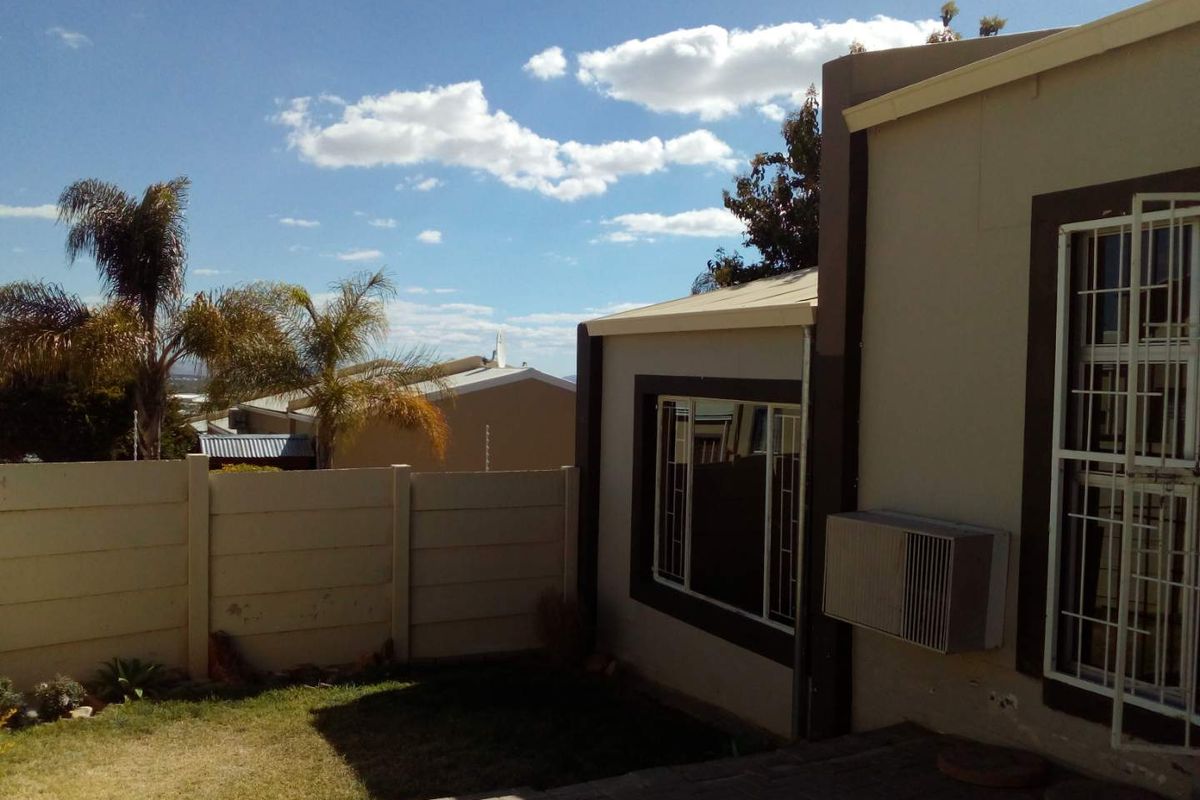 Rear exterior view of the townhouse featuring a small paved patio, lawn, precast boundary wall, and window-mounted air conditioning unit.