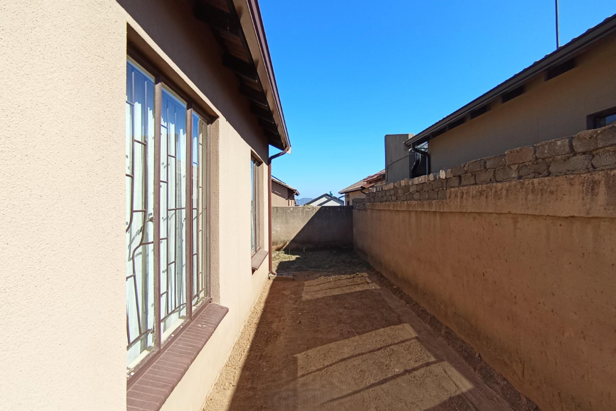 A narrow side exterior passage featuring a plastered house wall with security-barred windows and an unfinished boundary wall.