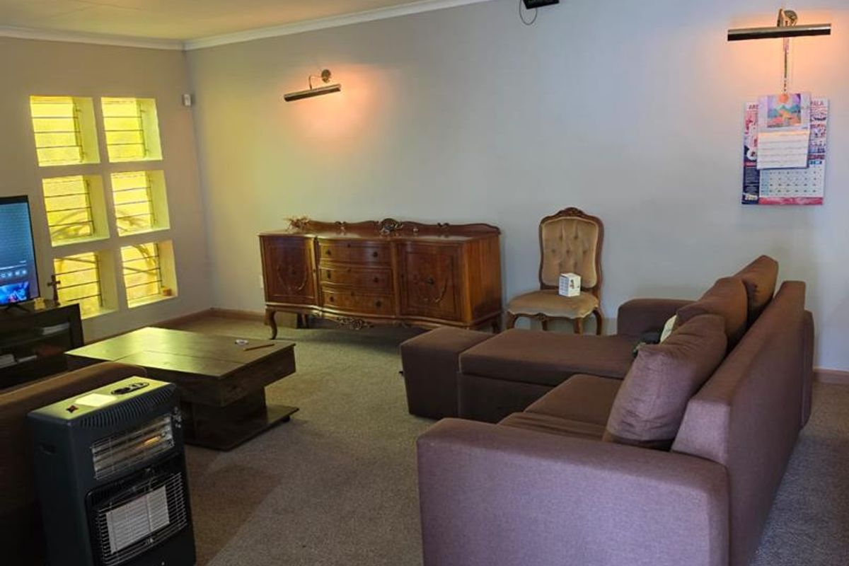 A spacious living room featuring carpeted flooring, louvered windows, and wall-mounted lighting, offering a functional and comfortable living space.