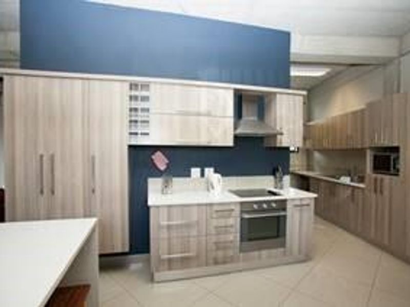 Modern office kitchen featuring light wood-grain cabinetry, stone countertops, integrated appliances including an oven and hob, and durable tiled flooring.