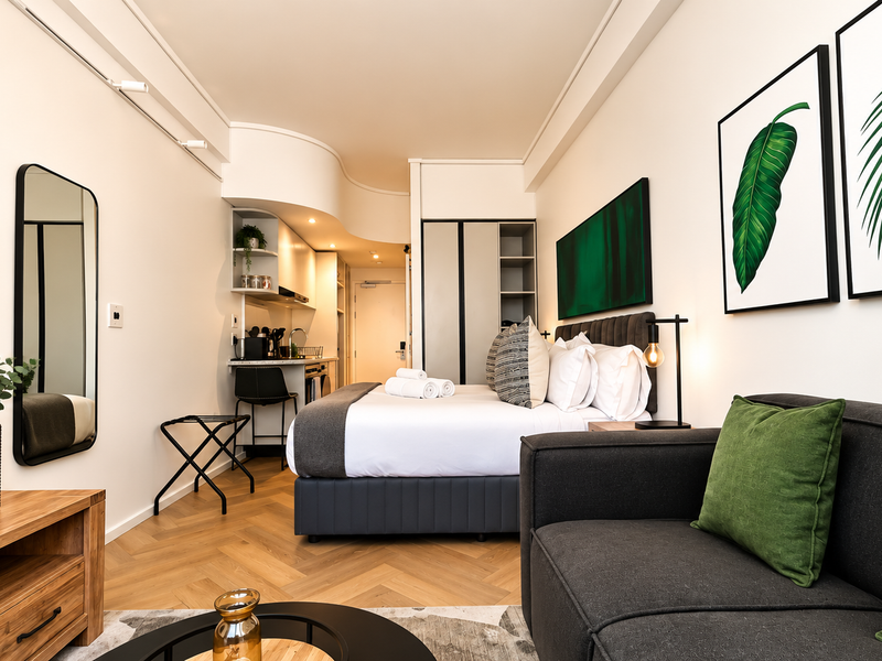 A modern open-plan studio apartment featuring herringbone timber flooring, a compact kitchenette, and built-in storage, finished with contemporary lighting and neutral decor.