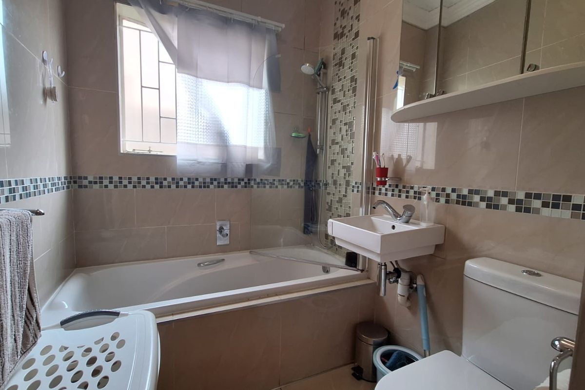 A compact bathroom featuring a bathtub with shower attachment, wall-mounted basin, toilet, and neutral wall tiling with a decorative mosaic accent border.