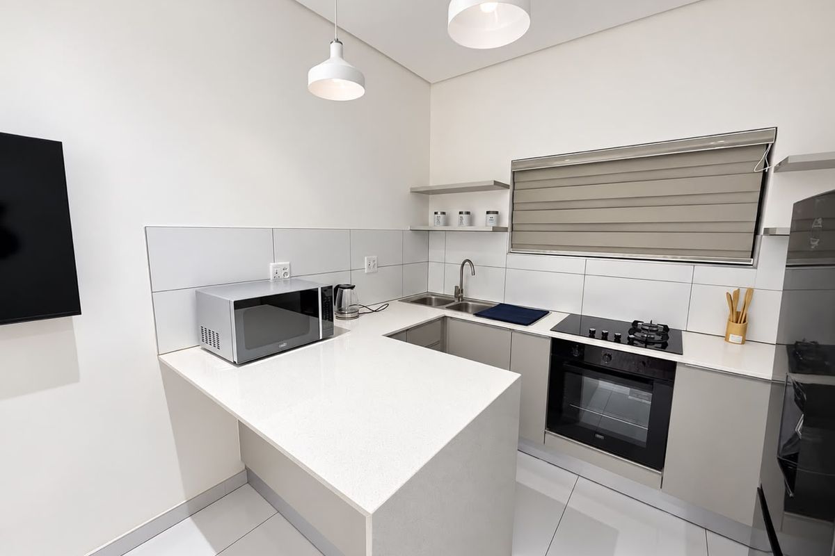 Modern kitchen featuring sleek quartz countertops, built-in oven, gas hob, stainless steel sink, and minimalist floating shelving with neutral tiled finishes.