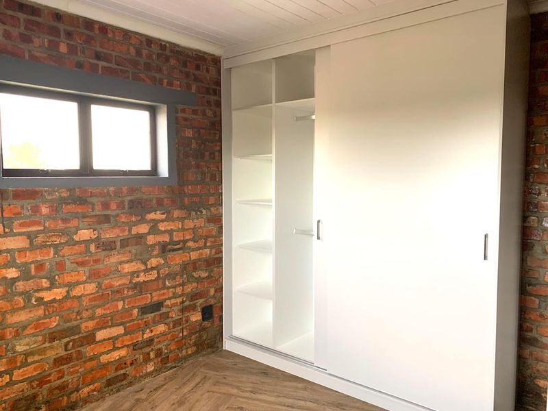 A bedroom featuring a modern white built-in wardrobe, exposed brick wall, and timber-look flooring, offering a clean and contemporary aesthetic.