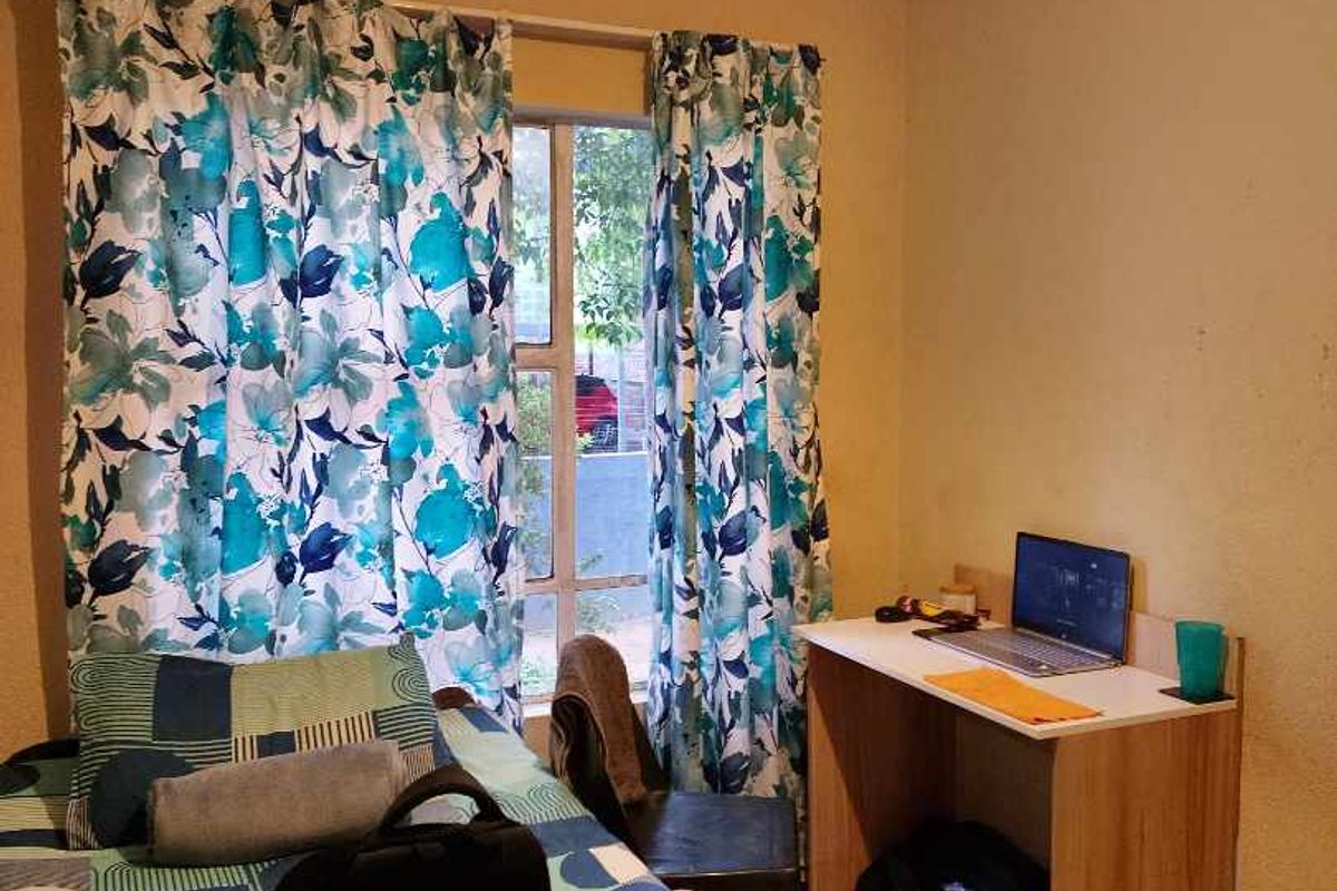 A compact bedroom featuring tiled flooring and a window with curtains, showing signs of wear on the wall surfaces.