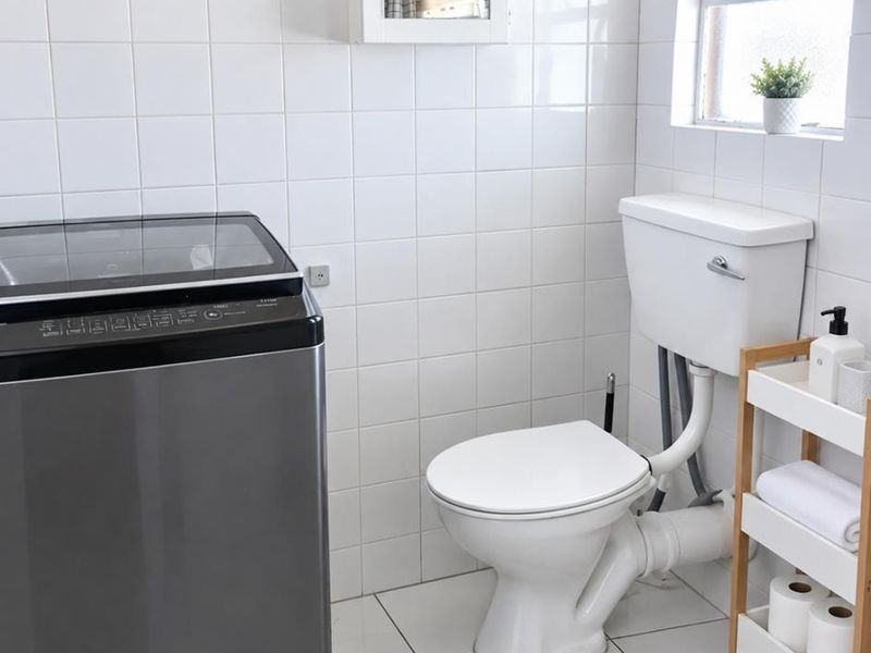 A functional laundry area featuring white wall and floor tiles, a toilet, and a wall-mounted storage cabinet.