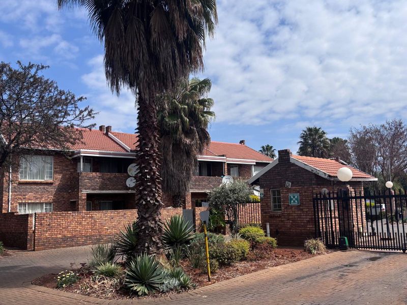 Exterior view of a face-brick apartment complex featuring a gated entrance, guard house, and paved driveway with established landscaping.