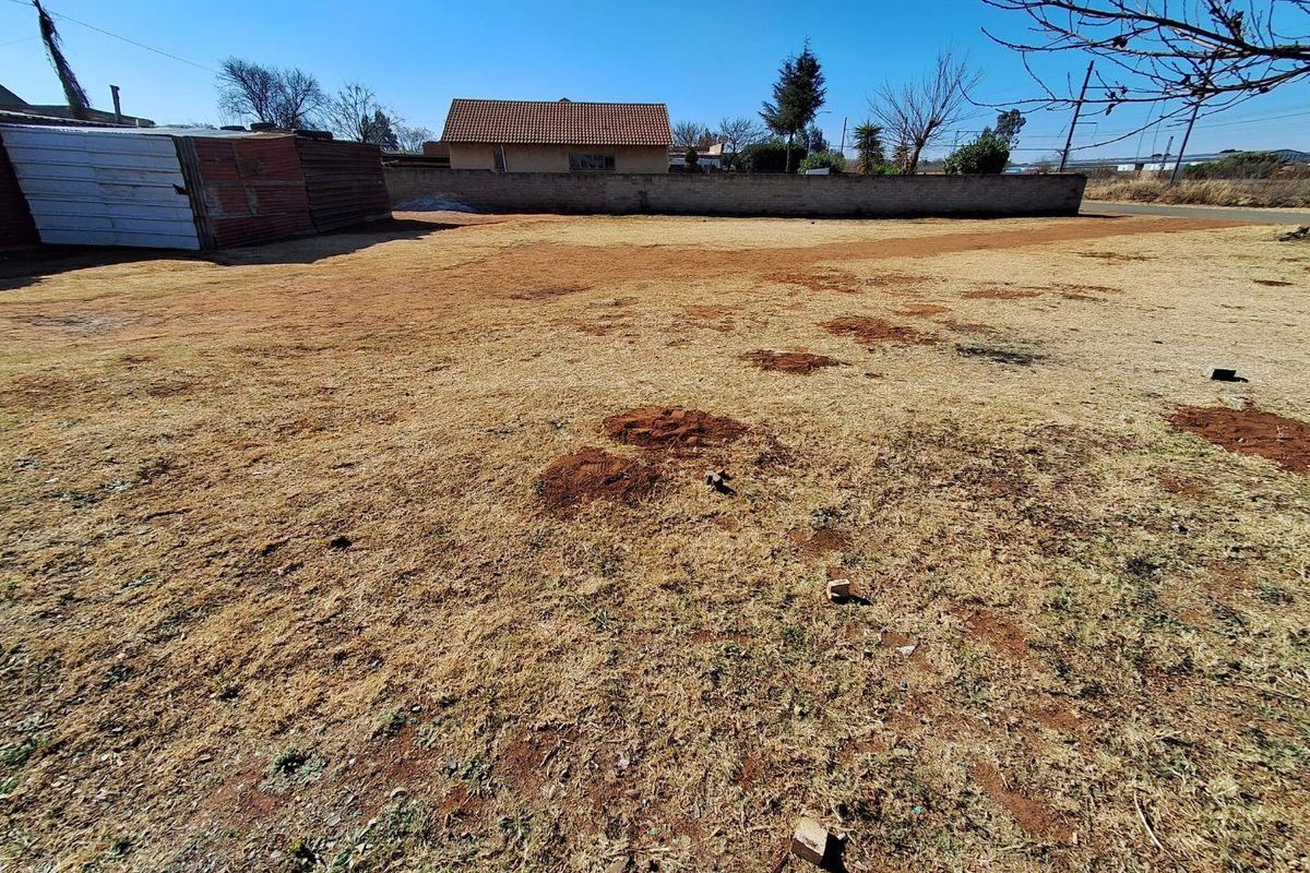 A vacant residential plot featuring dry grass, a perimeter boundary wall, and a small corrugated metal storage structure.