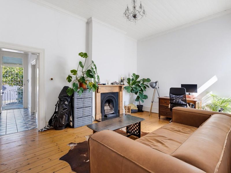 A bright living room featuring original timber flooring, a classic fireplace, and high ceilings with decorative cornicing, leading to an entrance hallway.
