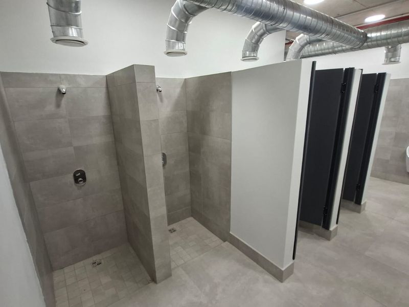 Modern industrial-style communal bathroom featuring tiled shower stalls, partitioned toilet cubicles, a wall-mounted urinal, and exposed overhead ventilation ducting.
