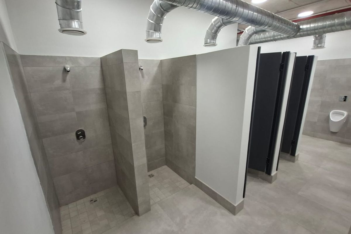 Modern industrial-style communal bathroom featuring tiled shower stalls, partitioned toilet cubicles, a wall-mounted urinal, and exposed overhead ventilation ducting.