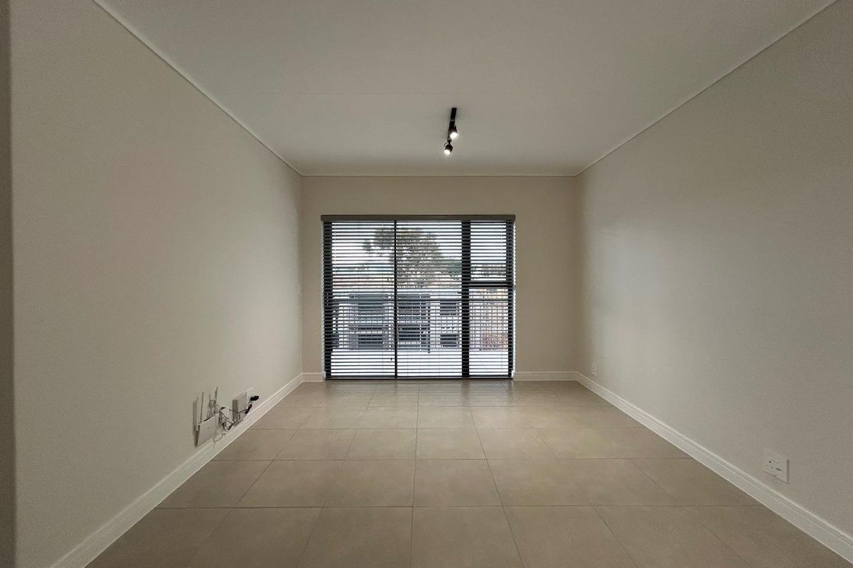 A bright, empty living area featuring neutral floor tiles, clean wall finishes, and a large window with fitted blinds.