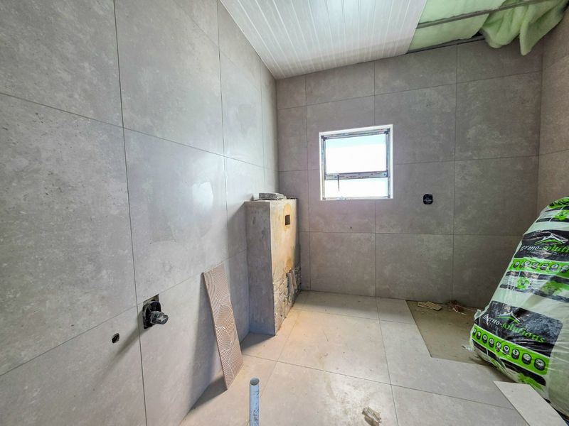 An unfinished bathroom space featuring large format grey wall and floor tiles, with plumbing rough-ins and exposed ceiling insulation ready for final installation.