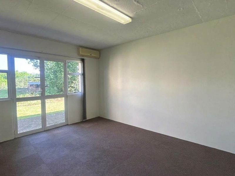 A bright, empty office space featuring carpeted flooring, a wall-mounted air conditioning unit, and a sliding glass door providing access to the exterior.