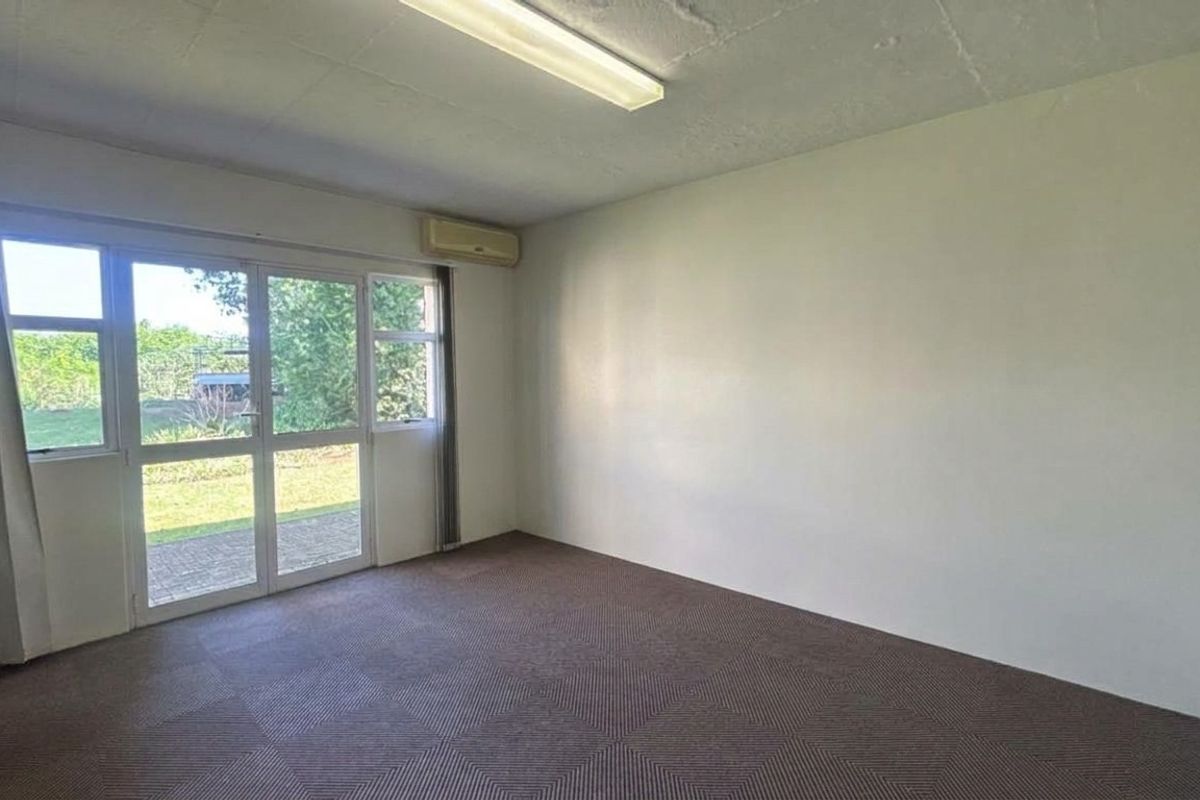 A bright, empty office space featuring carpeted flooring, a wall-mounted air conditioning unit, and a sliding glass door providing access to the exterior.