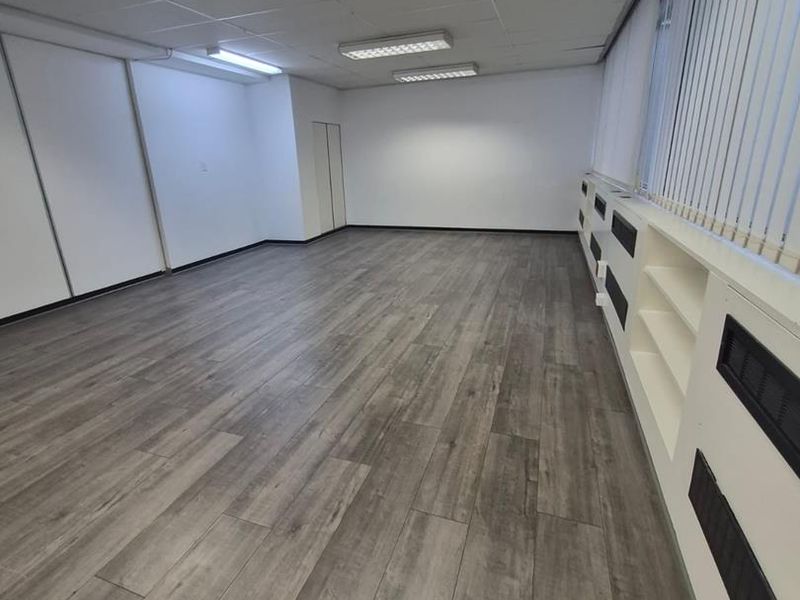 A spacious, empty office area featuring modern laminate flooring, suspended ceiling lighting, and built-in shelving units along the window wall.