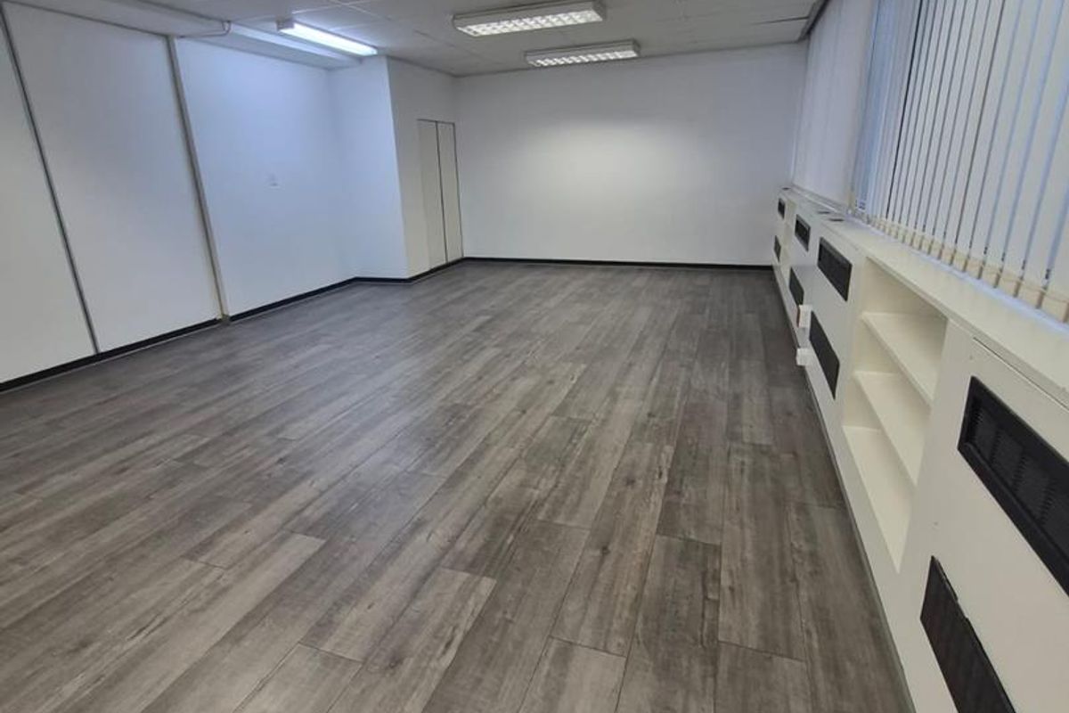 A spacious, empty office area featuring modern laminate flooring, suspended ceiling lighting, and built-in shelving units along the window wall.