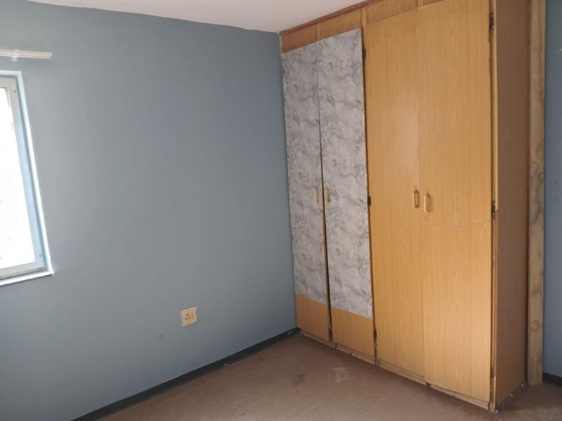 A bedroom featuring built-in cupboards with mismatched doors, blue painted walls, and a window, offering a blank canvas for renovation.