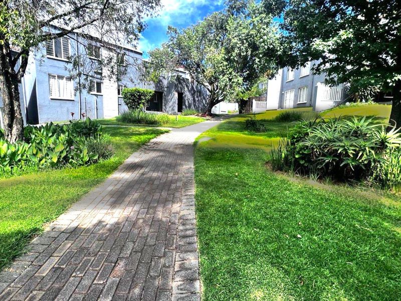 A view of the apartment complex grounds featuring a paved walkway winding through a well-maintained garden with mature trees and building facades.
