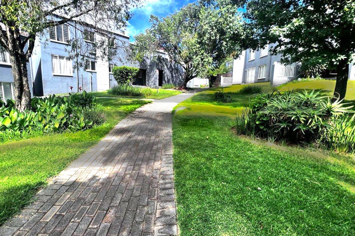 A view of the apartment complex grounds featuring a paved walkway winding through a well-maintained garden with mature trees and building facades.