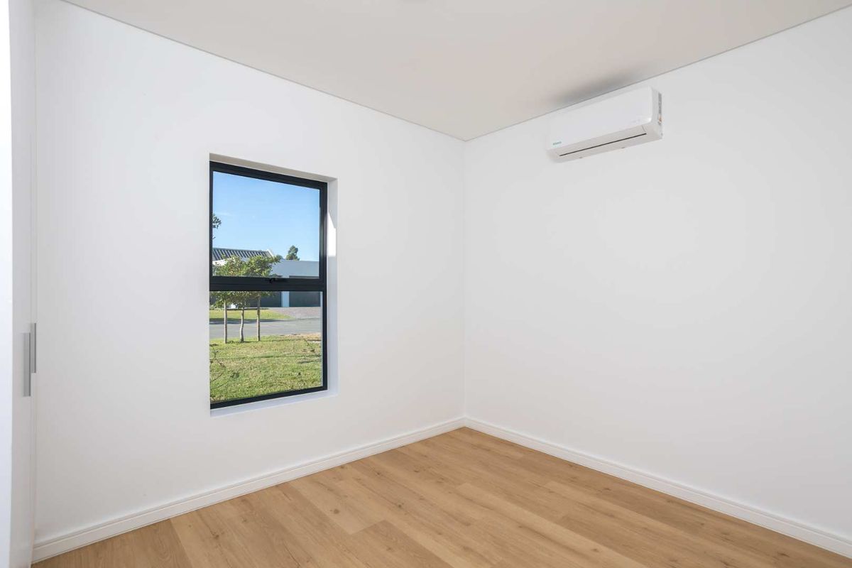 A bright, modern bedroom featuring light wood-look flooring, a wall-mounted air conditioning unit, and a window with a view of the exterior.