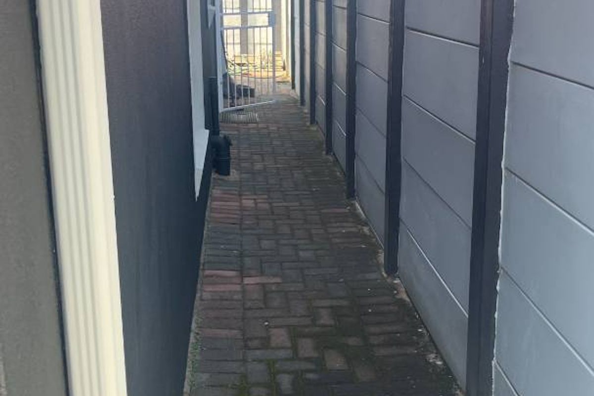 A narrow side passage featuring paved flooring, a boundary wall, and a gutter downpipe along the exterior of the house.