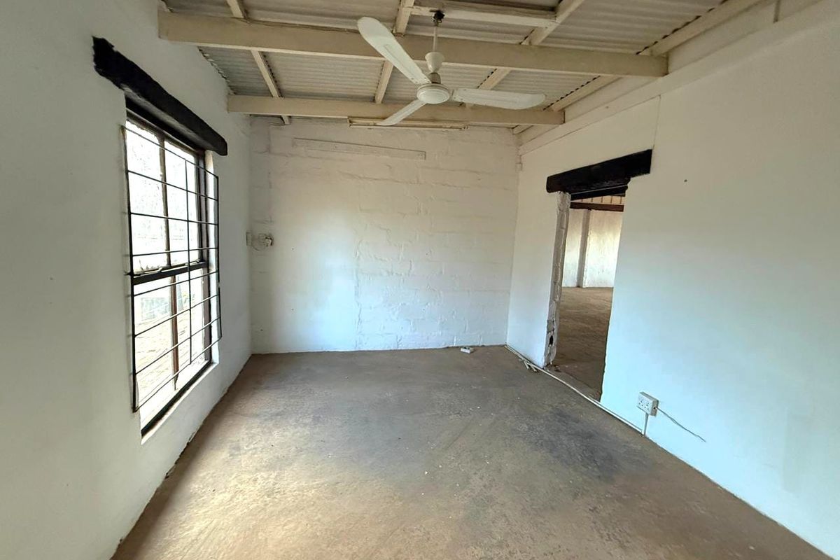 An empty, unfinished bedroom space featuring exposed ceiling beams, a ceiling fan, and security bars on the window, offering a blank canvas for renovation.