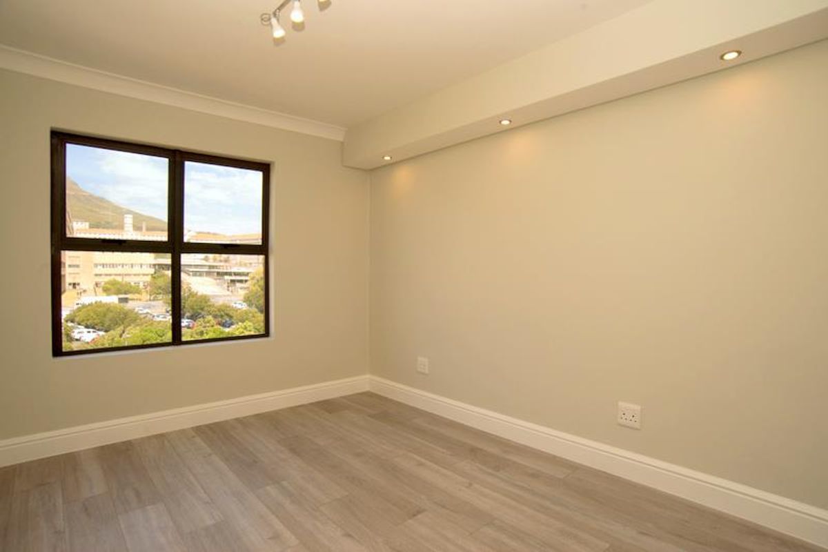 A bright, empty bedroom featuring modern laminate flooring, recessed downlighting, and a large window with views of the surrounding area.