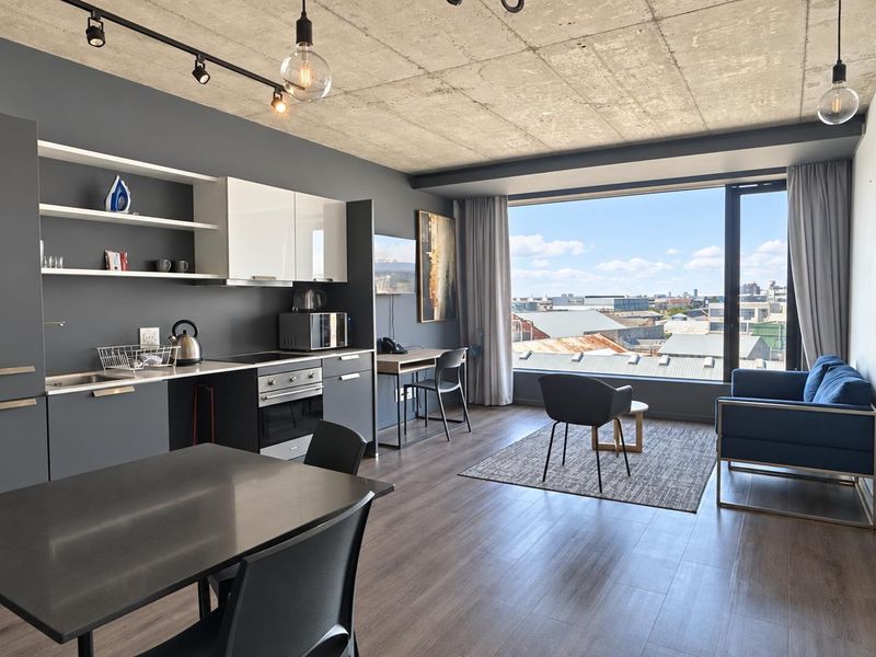 Modern open-plan apartment featuring an exposed concrete ceiling, sleek kitchen cabinetry with built-in appliances, and expansive windows offering panoramic urban views.