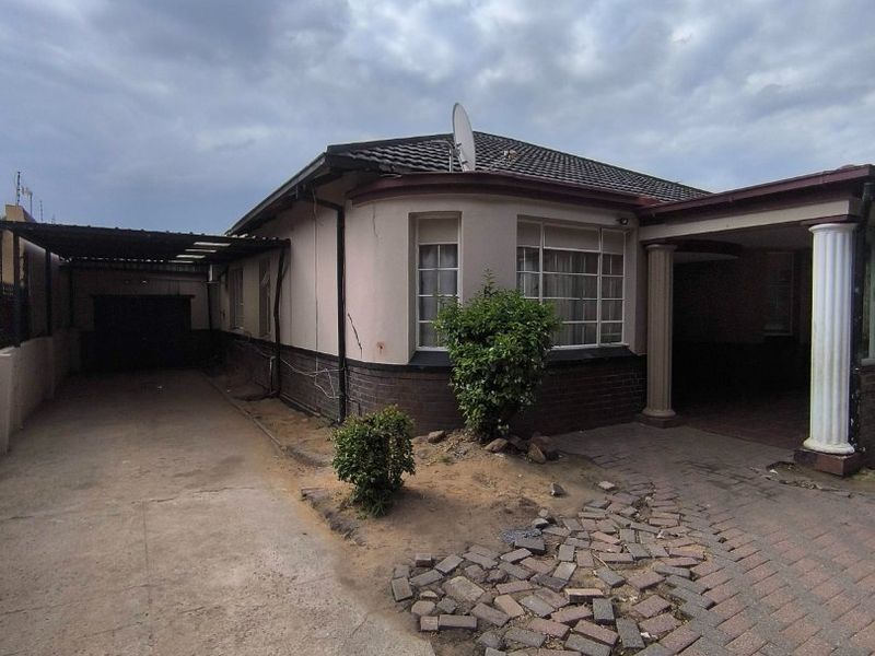 Front exterior view of a single-story house featuring a paved driveway, carport, and decorative columns, showing signs of wear on the exterior finishes.