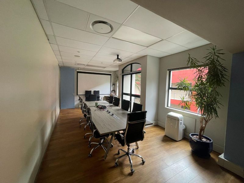 A functional office boardroom featuring laminate flooring, a suspended ceiling with recessed lighting, a projector screen, and large windows providing natural light.