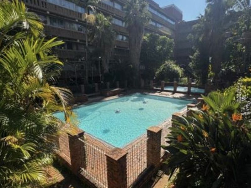 A communal swimming pool area featuring brick paving, metal fencing, and established landscaping within an apartment complex setting.