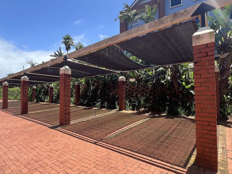 A covered parking area featuring brick pillars and shade cloth roofing, situated on a paved driveway with established garden landscaping.