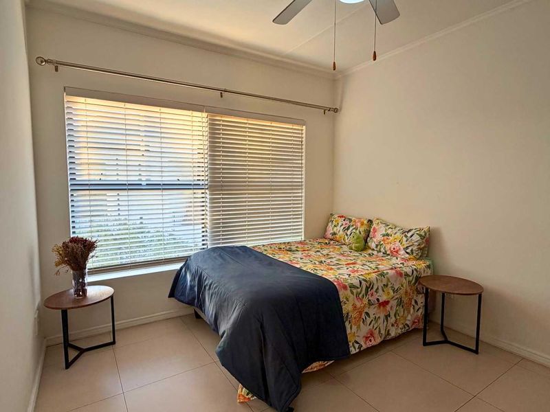 A functional bedroom featuring tiled flooring, a ceiling fan, and large windows fitted with venetian blinds for natural light control.