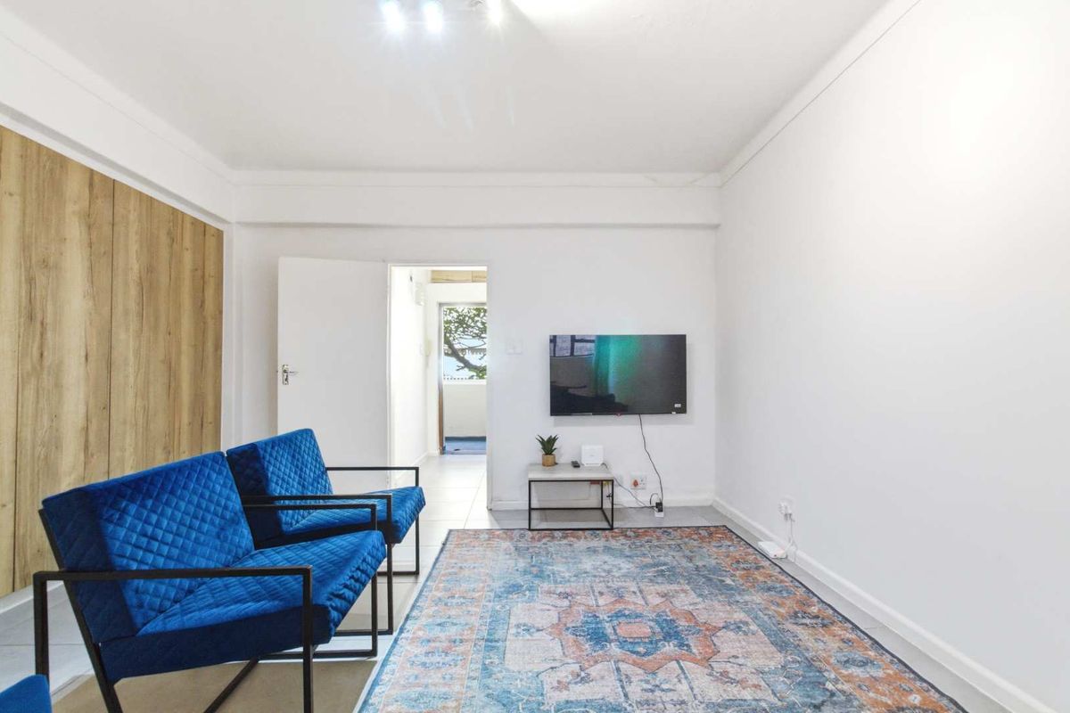 A bright and neutral living room featuring tiled flooring, a wood-look feature wall panel, and direct access to an adjacent hallway.