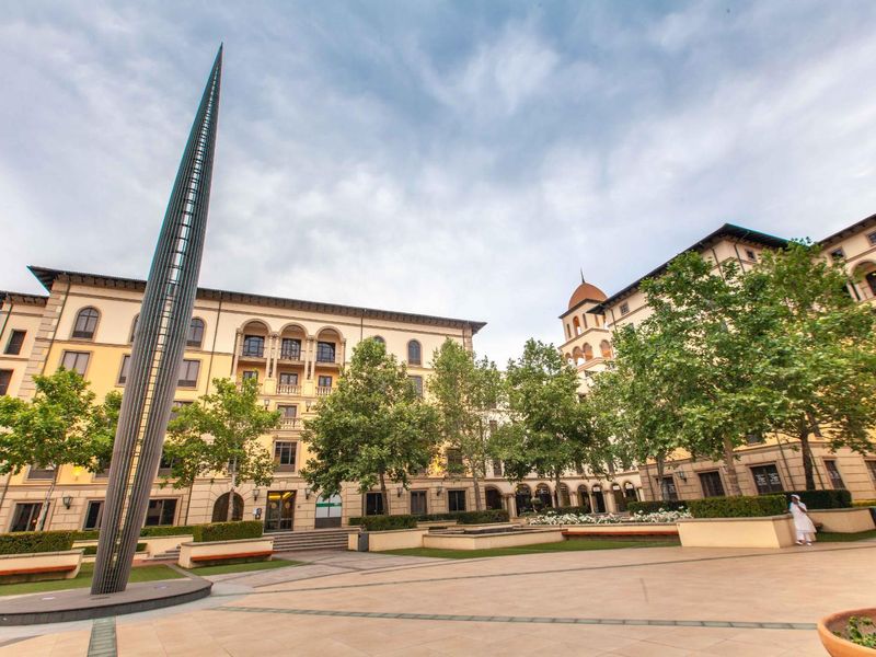 A well-maintained office complex courtyard featuring paved walkways, mature trees, landscaped garden beds, and a prominent sculptural spire.