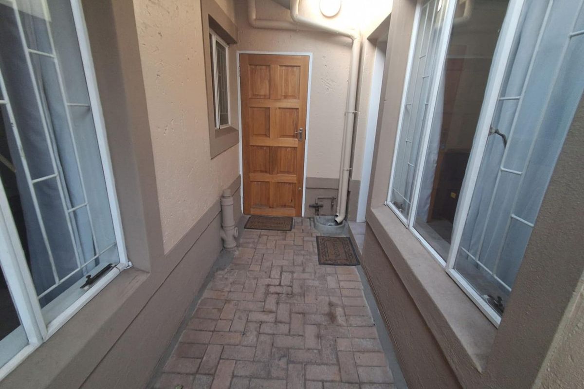 A narrow paved exterior walkway between townhouse walls featuring a timber door, downpipe infrastructure, and security-barred windows.