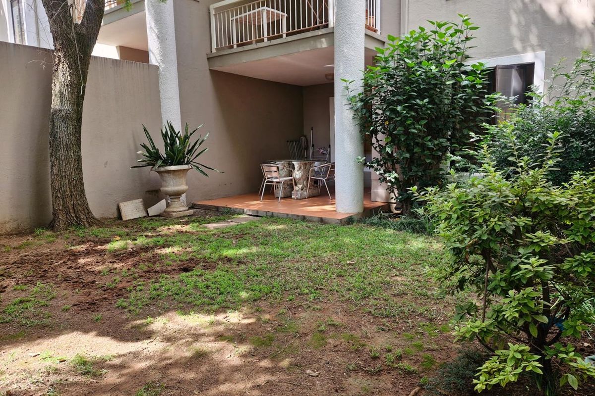 A private ground-floor patio area featuring tiled flooring and structural columns, overlooking a small garden space with established trees and shrubs.