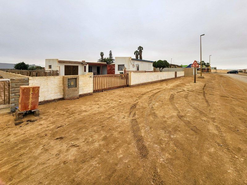 Street view of the property showing the exterior boundary wall, sliding gate entrance, and unpaved road access.