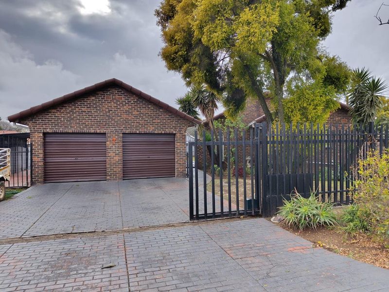 Front view of a brick house featuring a double garage, paved driveway, and secure metal perimeter fencing.
