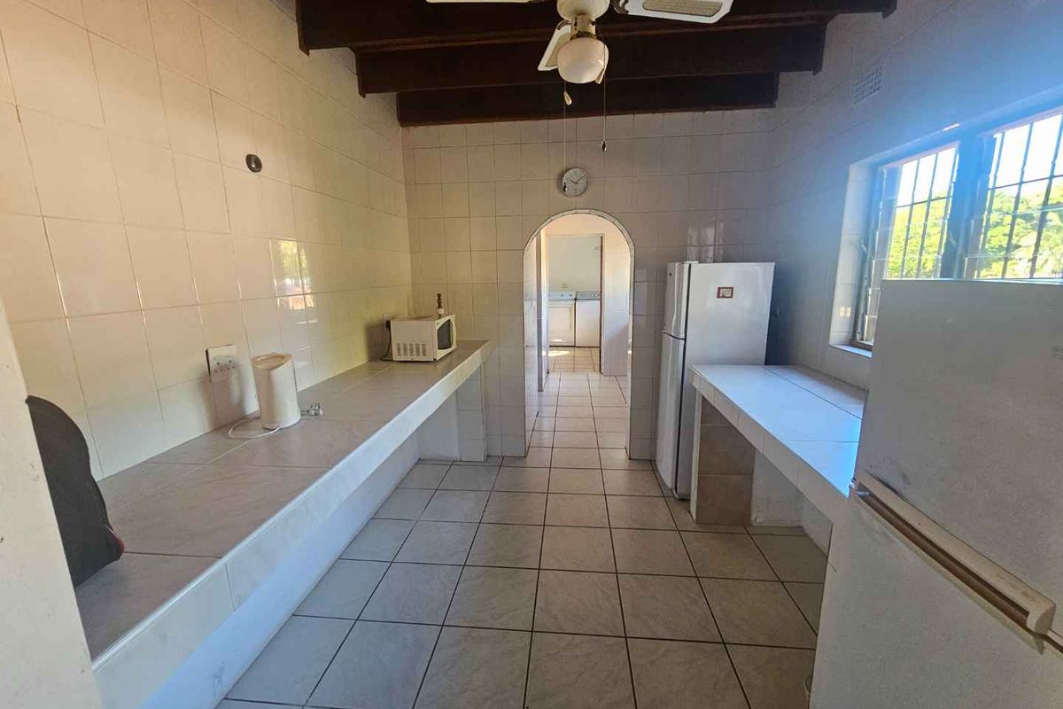 A functional kitchen featuring tiled countertops, exposed timber ceiling beams, and a ceiling fan, with an arched doorway leading to an adjacent utility area.