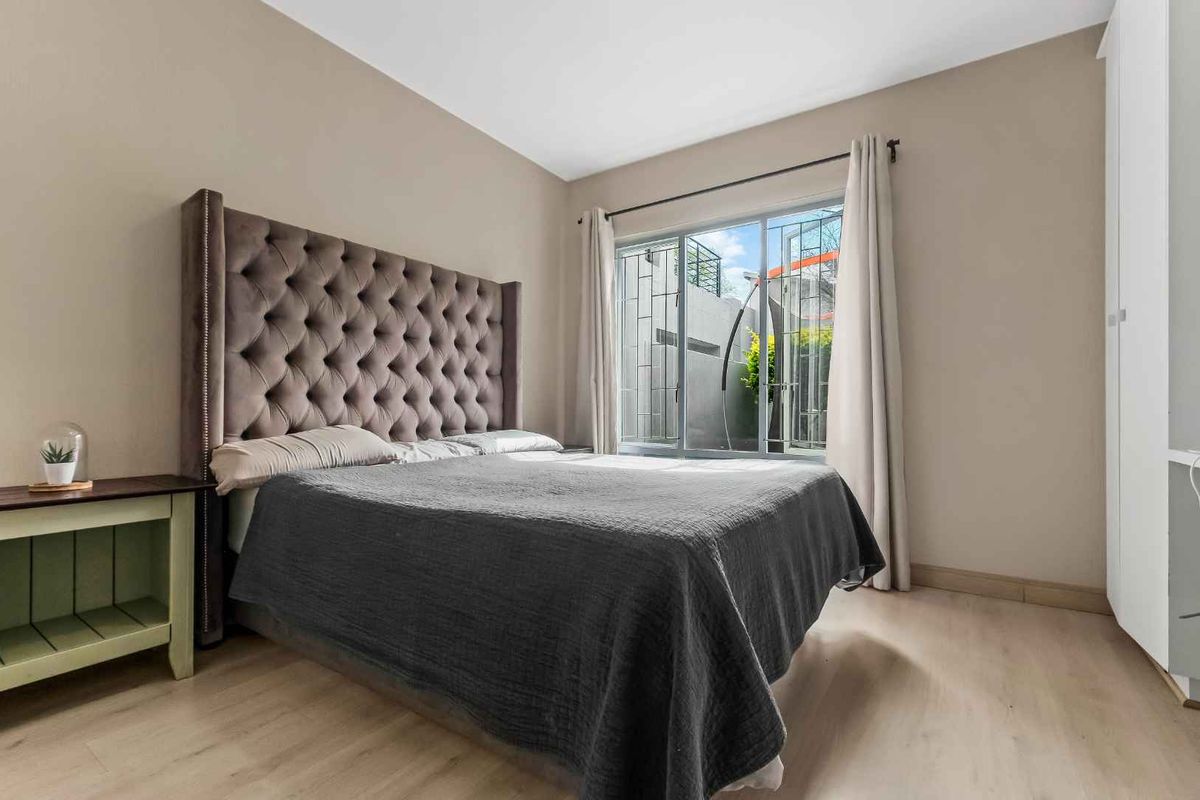 A well-lit bedroom featuring light wood-look flooring, neutral wall tones, and a large window with security bars, providing a comfortable and functional living space.