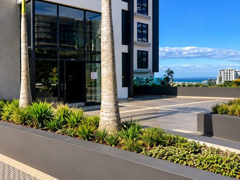 Modern building exterior featuring a glass entrance, paved driveway, and landscaped planter boxes with a clear view of the surrounding area.