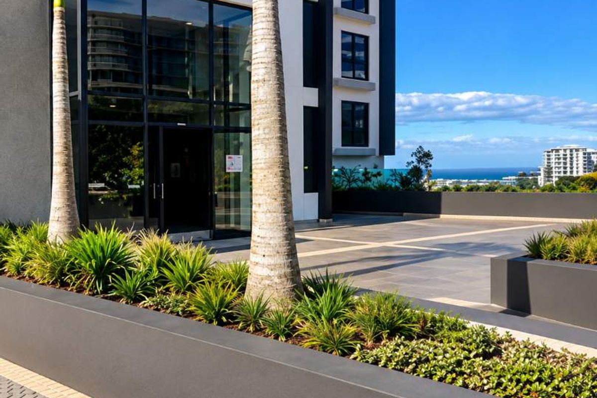 Modern building exterior featuring a glass entrance, paved driveway, and landscaped planter boxes with a clear view of the surrounding area.