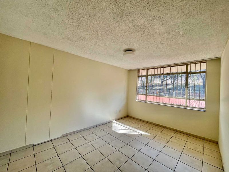 An empty bedroom featuring tiled flooring, a textured ceiling, and a large window with security bars, offering a blank canvas for renovation.