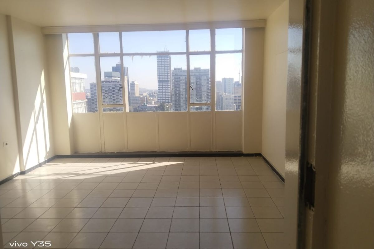 An empty, sunlit living area featuring large metal-framed windows with expansive city views and neutral tiled flooring throughout.